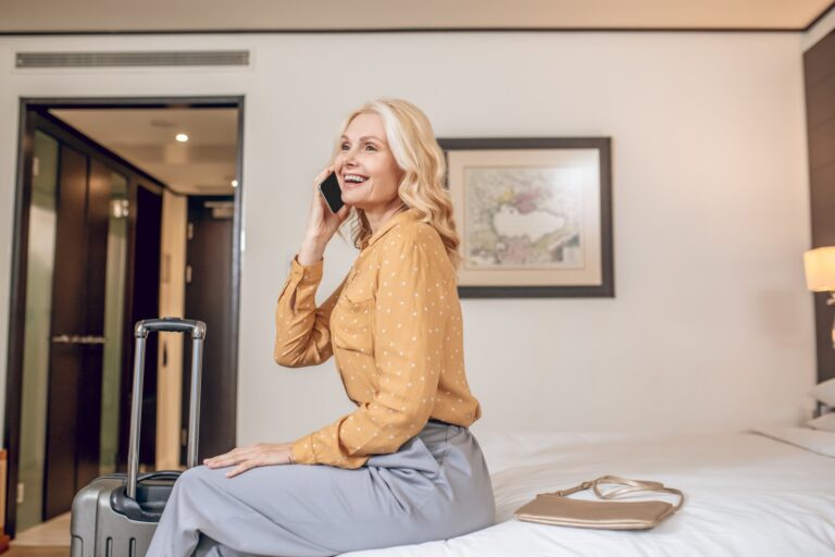 Smiling woman sitting on a hotel bed beside her suitcase, on the phone
