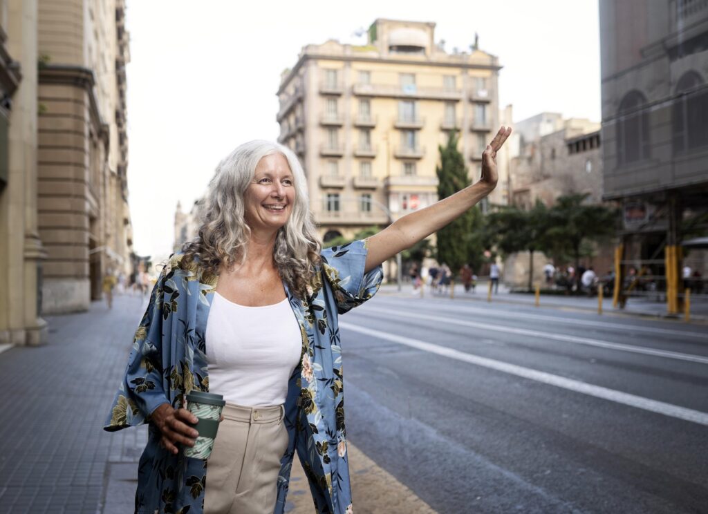 Smiling woman with long gray hair waving on a city street, coffee cup in hand