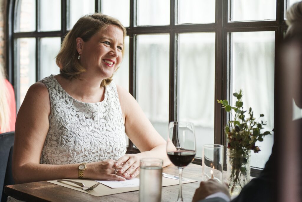 Woman dining on her own at a restaurant table by the window, smiling, with a glass of wine