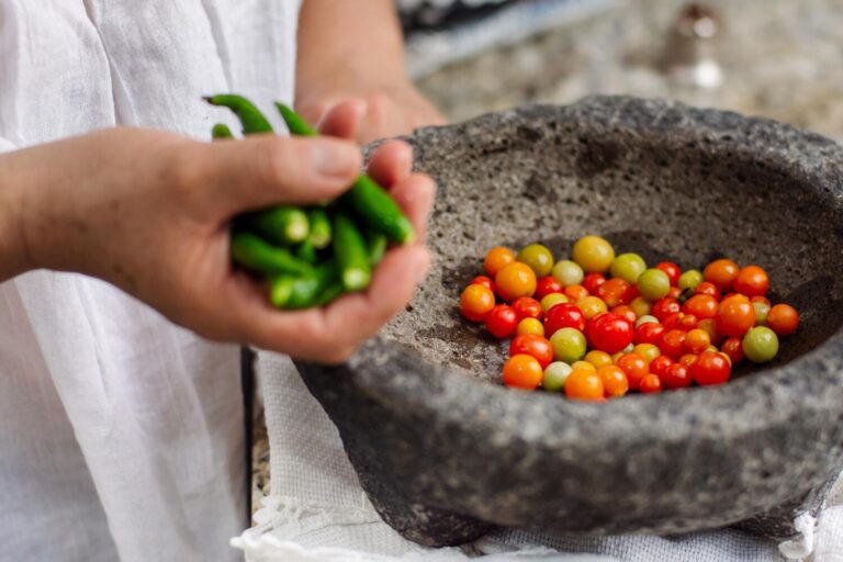Cook adding green chiles to tomatoes in a stone molcajete to make salsa